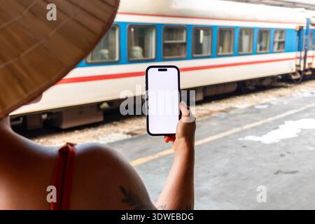 Tourist in non la holding smartphone with blank white screen on a Hanoi train platform, back view capturing travel, culture and modern technology meet Stock Photo