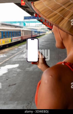 Tourist wearing a traditional conical hat waiting on a train station platform, holding a modern frameless smartphone with a blank white screen, travel Stock Photo