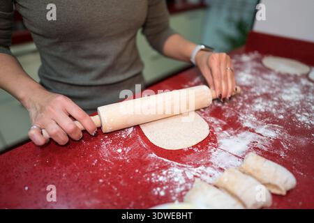 A person rolls out bread dough with a wooden rolling pin on a red countertop, surrounded by flour and folded dough pieces, emphasizing homemade baking Stock Photo