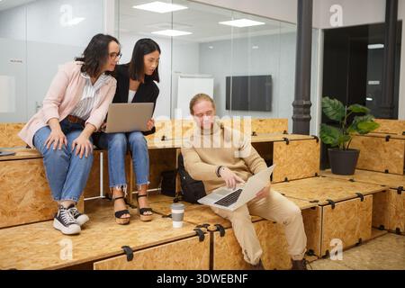 A diverse group of professionals collaborating in a modern coworking space. They're engaged in a casual business discussion, using laptops to brainsto Stock Photo