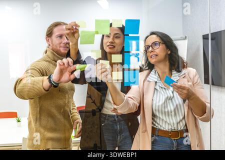 A diverse team collaborates in a modern office, engaged in a discussion. They work together in a lively coworking space, using sticky notes for brains Stock Photo
