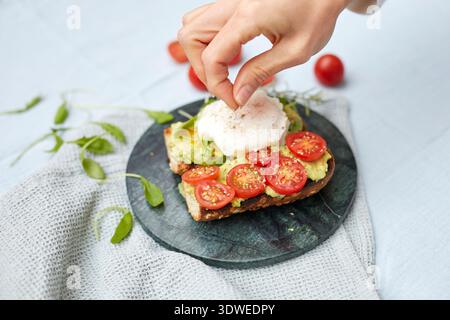 A hand sprinkles seasoning over a poached egg on avocado toast. The dish is beautifully garnished with sliced cherry tomatoes, creating a vibrant brea Stock Photo