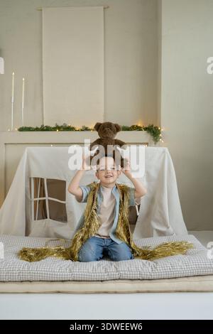 A joyful boy sits with a teddy bear on his head, surrounded by festive Christmas decorations and lights. The scene captures the spirit of childhood de Stock Photo