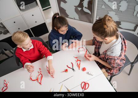 A group of kids and an adult engaged in a festive craft session, creating Christmas decorations using plasticine and ribbons. The table is filled with Stock Photo