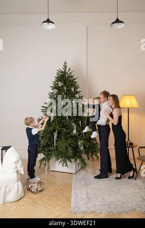 A joyful family gathers around a beautifully adorned Christmas tree in a cozy living room, engaging in the beloved tradition of decorating together fo Stock Photo