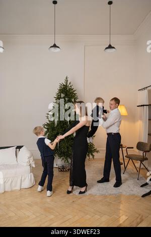 A joyful family celebrating New Year together at home. Parents and children enjoy time around a beautifully decorated holiday tree, creating festive m Stock Photo
