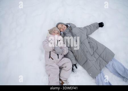 Two sisters enjoy the snowy winter day in the park, lying on their backs in warm clothing and sharing smiles. Their winter outfits keep them snug and Stock Photo