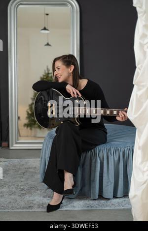 A woman sits on a bed with a black electric guitar, smiling. The modern room features a large mirror, gray walls, a blue bed cover, and pendant lights Stock Photo