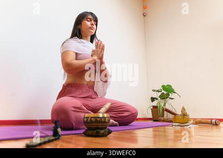 Woman practicing yoga in a serene indoor setting, sitting cross-legged on a purple mat. Surrounded by calming elements like incense and plants, she is Stock Photo