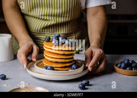 A woman arranges a delicious breakfast plate with a stack of fluffy pancakes topped with fresh blueberries. The scene is set on a modern, minimalist k Stock Photo