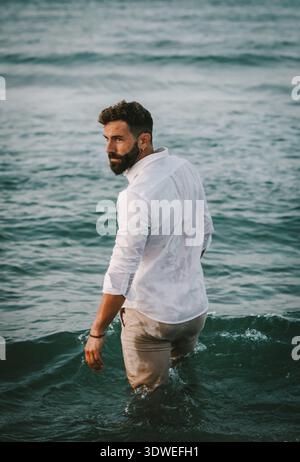A confident man with a beard stands waist-deep in the ocean, wearing a white shirt. The serene sea setting reflects a healthy lifestyle, capturing the Stock Photo