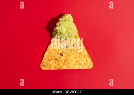 A single Mexican nacho chip is generously topped with creamy guacamole on a vibrant red background. The bright colors highlight the appetizing texture Stock Photo