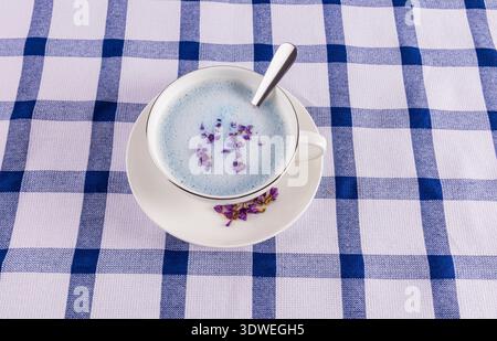 A delicate blue matcha latte in a white cup with a spoon, topped with purple flower petals. Served on a white saucer, placed on a blue and white check Stock Photo