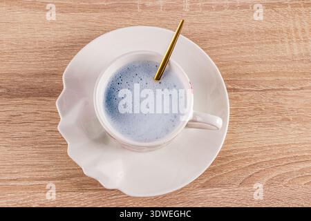 A frothy blue matcha latte fills a decorative white cup with a gold spoon resting inside, set on a wooden table. The vibrant blue color contrasts beau Stock Photo