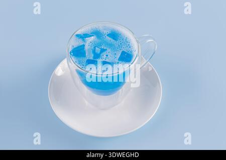 A clear glass cup filled with a vibrant blue matcha beverage, served on a white saucer. The blue liquid includes ice cubes, set against a matching blu Stock Photo