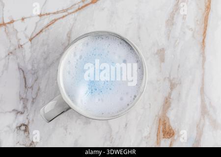 A frothy blue matcha latte in a cup sits atop a textured marble surface. The vibrant blue hue contrasts elegantly with the neutral background, offerin Stock Photo