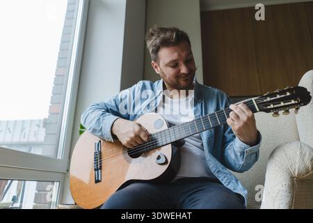 A young man casually plays an acoustic guitar in his cozy home environment. He is focused and relaxed, enjoying the calming atmosphere by the window o Stock Photo