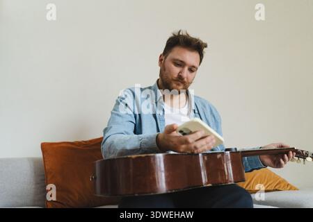A young man with smartphone sits comfortably on a couch at home, playing an acoustic guitar. He appears focused and relaxed, enjoying his time immerse Stock Photo