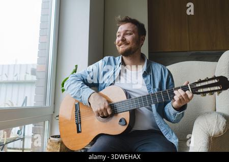 A young man sits comfortably on an armchair by the window, playing an acoustic guitar. He appears relaxed and content, enjoying a peaceful moment in h Stock Photo