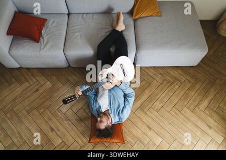 A young man lies comfortably on the floor at home, playing an acoustic guitar. Surrounded by soft pillows and casual attire, he enjoys a moment of mus Stock Photo