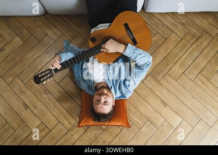 A young man lies comfortably on the floor, strumming an acoustic guitar. He is relaxed in a casual setting, showcasing an intimate moment of musical e Stock Photo