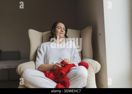 A woman sits contentedly in an armchair, immersed in the calming hobby of knitting a vibrant red sweater. Her relaxed posture reflects the joy of crea Stock Photo