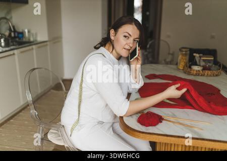 A woman focuses on her knitting project, carefully holding a red sweater while talking on her phone. Her passion for this hobby is evident in her thou Stock Photo