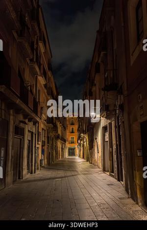 A serene, narrow medieval street in Tarragona, Spain is softly illuminated by warm streetlights at night, casting shadows on the cobblestones and show Stock Photo