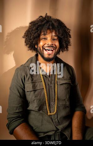 A cheerful man with an afro hairstyle is captured smiling brightly in a studio setting. He wears a casual shirt and a gold chain, exuding warmth and s Stock Photo
