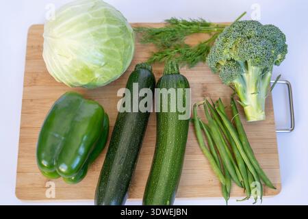 A high-angle flat lay shot of organic green vegetables arranged on a bamboo cutting board. The set includes a head of cabbage, two zucchinis, a bell p Stock Photo