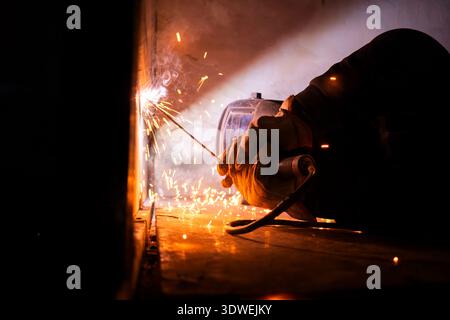 Unrecognizable man welds metal with sparks flying, using a protective shield and gloves. The glow of the welding process highlights the intensity of t Stock Photo