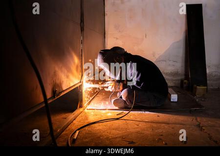 Unrecognizable man engaged in welding work, wearing a protective mask. Sparks fly as he welds metal in a dimly lit industrial setting, focused and ski Stock Photo