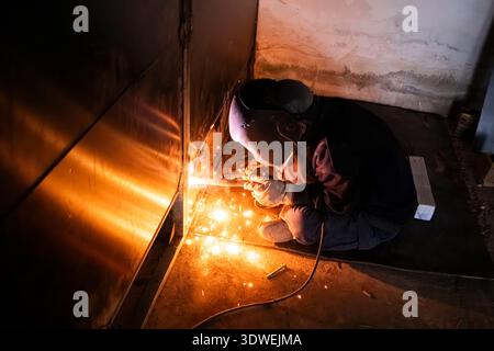 Unrecognizable man welding with protective mask, crouching in a dimly lit workshop environment surrounded by sparks and metal, emphasizing safety and Stock Photo
