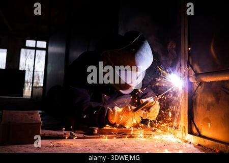 Unrecognizable man welding indoors with protective mask and gloves, creating bright sparks. The dimly-lit workshop emphasizes the intense work environ Stock Photo