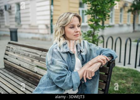 A thoughtful woman with blonde hair sits on a wooden bench wearing a denim jacket. The street backdrop features buildings and greenery, creating a pea Stock Photo