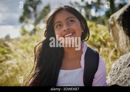 Cheerful latin american elementary school student smiling and looking away, enjoying a field trip in nature, surrounded by vibrant plants and trees on Stock Photo