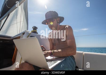 A man is working on a laptop under the sun on a sailboat in the Mediterranean Sea, combining leisure and technology for a modern maritime lifestyle. Stock Photo