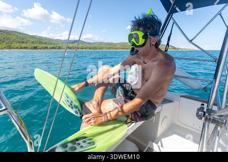 A man prepares for a snorkeling adventure on a sailboat, wearing flippers and a snorkel mask, surrounded by the calm, clear waters of the Mediterranea Stock Photo