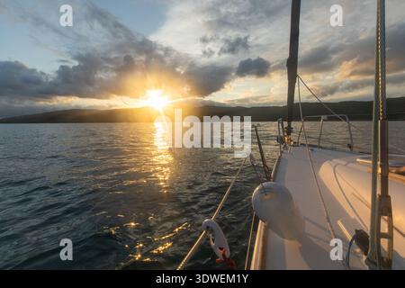 A serene scene of sailing in the Mediterranean at sunset, with the sun setting behind distant hills and casting a warm glow over the tranquil sea, cre Stock Photo
