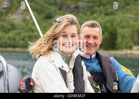 A couple smiles joyfully while sailing through Norway picturesque waters. The lush green landscape provides a serene backdrop, capturing the essence o Stock Photo