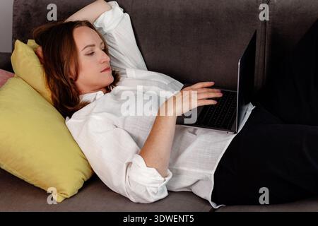 A woman reclines comfortably on a sofa at home, using a laptop for freelance work. She appears focused and relaxed in her casual setting, showcasing m Stock Photo