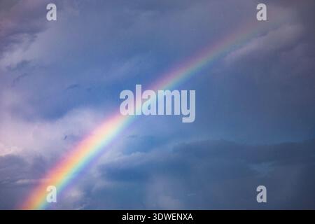 A vivid rainbow stretches across a dramatic cloudy sky, showcasing a spectrum of colors from red to violet. The contrast of light against dark creates Stock Photo