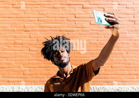 A man with an afro hairstyle takes a selfie against a sunlit orange brick wall. He is wearing a brown textured shirt and appears relaxed and confident Stock Photo