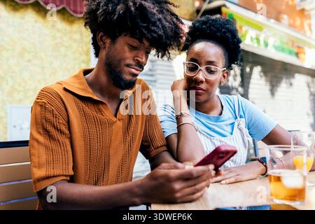 A young couple sits at an outdoor cafe, casually interacting while looking at a smartphone. They are enjoying their drinks, dressed in relaxed casual Stock Photo
