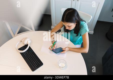 A girl diligently works on her homework at a white table, using a smartphone for assistance. She is in a modern home setting, surrounded by study mate Stock Photo