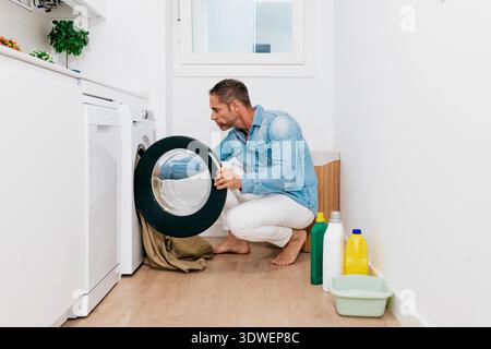 Man in a casual denim shirt doing laundry in a bright, modern utility room. He kneels by a front-loading washing machine, with cleaning supplies nearb Stock Photo