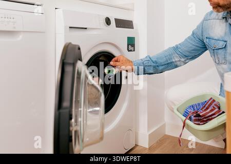 A man in a denim shirt kneels by a front-loading washing machine in a bright laundry room, loading detergent and clothes in a basket, focused on house Stock Photo