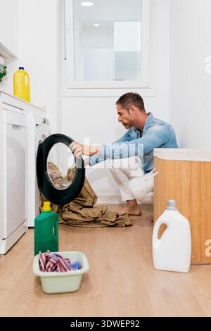 A man kneels in a modern laundry room loading clothes into a front-loading washing machine. Cleaning supplies are scattered on the wooden floor, addin Stock Photo