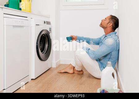 A man in casual attire sits on a wooden floor next to a washing machine, holding a cup of coffee. He appears relaxed, suggesting a break from house cl Stock Photo