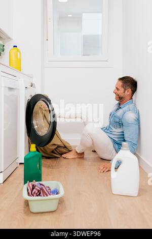 Smiling man sitting on the floor doing laundry at home. He's using a washing machine to clean clothes. Laundry detergent and basket nearby add to the Stock Photo
